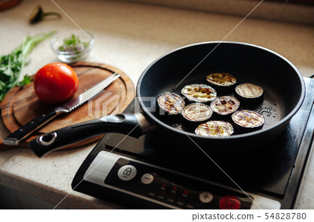 Eggplants are fried in a skillet. Vegetables on a cutting board in the background. 54828780