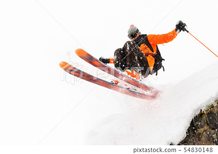 The athlete skier on a light background with a jump moves off the roof of a snow-covered hut with 54830148