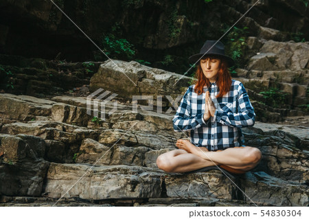 Beautiful girl with red hair in a hat and shirt meditating on rocks in a lotus position. Space for Beautiful girl with red hair in a hat and shirt meditating on rocks in a lotus position. Space for 54830304