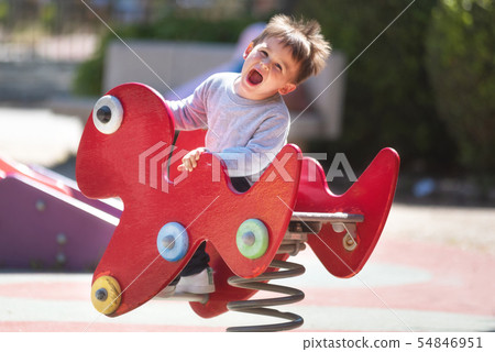 Cute happy baby boy with blond hair riding red spring rider or rocker on sunny summer day. 54846951