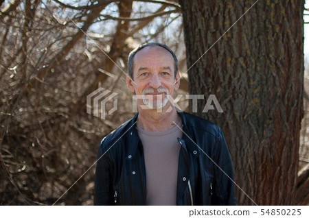 Senior handsome man wearing a dark leather jacket standing outdoors near the tree on blue sky Senior handsome man wearing a dark leather jacket standing outdoors near the tree on blue sky 54850225