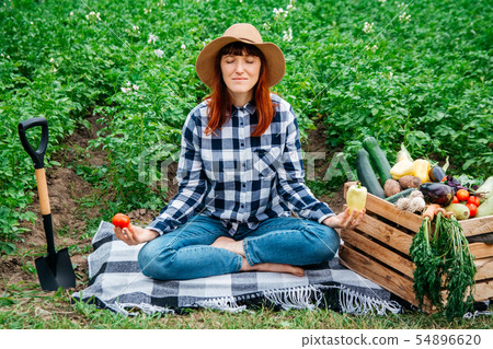 Beautiful female farmer meditating while sitting on a blanket near fresh organic vegetables in a 54896620