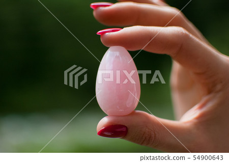Close up photo of female hand holding a yoni egg. Rose quartz crystal egg on river background Close up photo of female hand holding a yoni egg. Rose quartz crystal egg on river background 54900643