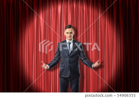 Young serious businessman with palms up on red stage curtains background 54906355