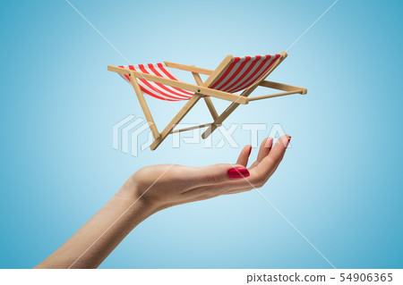 Side closeup of woman's hand facing up and levitating small striped chaise-longue on light blue 54906365