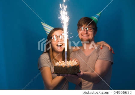Funny nerd man and woman are wearing holidays caps and glasses holding birthday cake with candles Funny nerd man and woman are wearing holidays caps and glasses holding birthday cake with candles 54906653