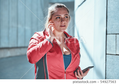 Attractive plus size woman in headphones holding smart phone and listening music while standing Attractive plus size woman in headphones holding smart phone and listening music while standing 54927563