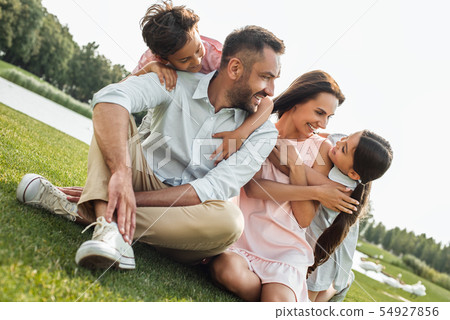 Summertime. Happy young family of four smiling and having fun while sitting on grass in park 54927856