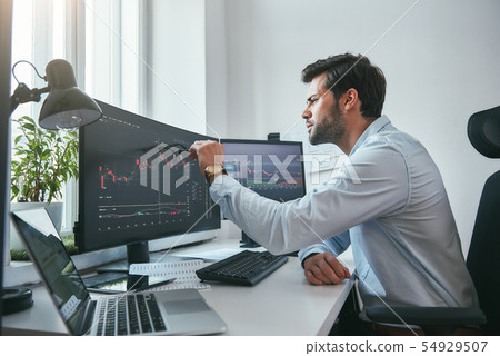 Analyzing data. Thoughtful young trader is pointing at graphs on computer screen while working in 54929507