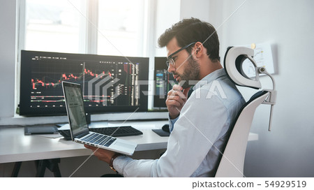 Busy day. Professional male trader wearing eyeglasses analyzing financial market via laptop while 54929519