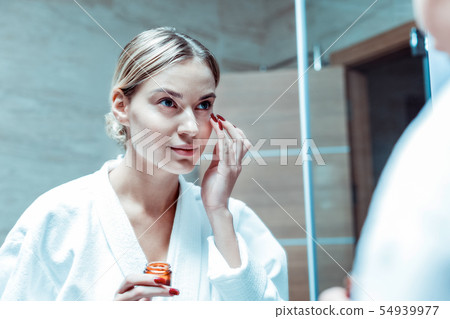 Focused blonde woman nourishing her eye area with cream Focused blonde woman nourishing her eye area with cream 54939977