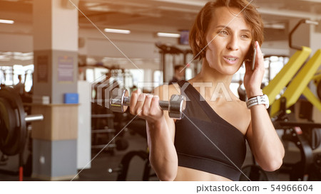 Mature woman is making biceps exercise with dumbbells in gym and speaking phone. Mature woman is making biceps exercise with dumbbells in gym and speaking phone. 54966604