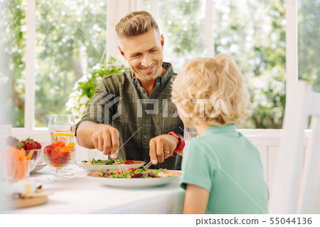 Handsome husband smiling while cutting salad for his curly son Handsome husband smiling while cutting salad for his curly son 55044136