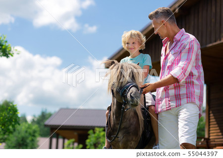 Dark-eyed curly son feeling happy riding horse for the first time Dark-eyed curly son feeling happy riding horse for the first time 55044397