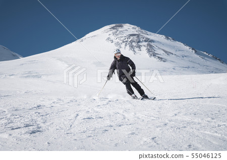 A bearded mature aged male skier in a black ski suit descends along the snowy slope of a ski resort 55046125