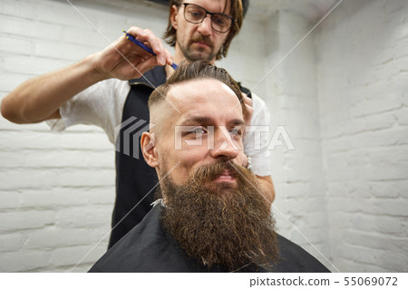 Master cuts hair and beard of men in the barbershop, hairdresser makes hairstyle for a young man Master cuts hair and beard of men in the barbershop, hairdresser makes hairstyle for a young man 55069072