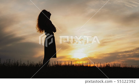 Silhouette of a young woman standing in dry grass field on sunset Silhouette of a young woman standing in dry grass field on sunset 55122392
