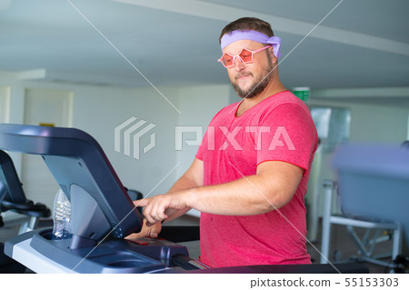 Funny fat male in pink glasses and in a pink t-shirt is engaged on a treadmill in the gym depicting 55153303