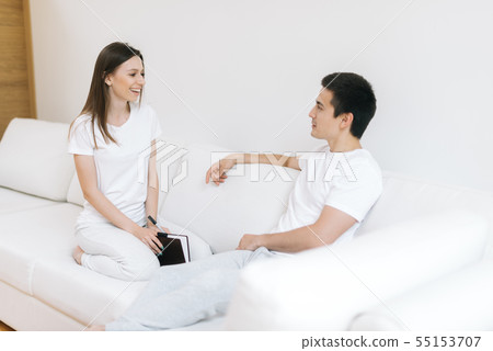 Young happy couple sitting on white sofa, smiling at each other Young happy couple sitting on white sofa, smiling at each other 55153707