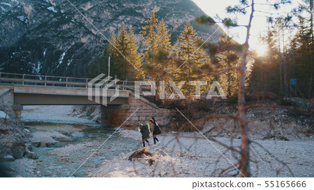 Two young women crossing the rocky river with the backpacks and guitar under a bridge in the 55165666