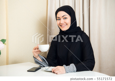 Attractive young female muslim holding coffee mug cup sitting at home and smiling to the camera Attractive young female muslim holding coffee mug cup sitting at home and smiling to the camera 55171572