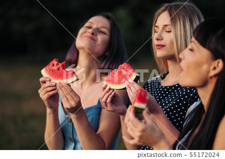 Three young attractive women friends in blue dresses at sunset are eating watermelon and smiling. 55172024