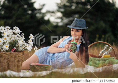 A young beautiful woman in hat and dress is drinking lemonade from a glass jar, sitting on plaid on A young beautiful woman in hat and dress is drinking lemonade from a glass jar, sitting on plaid on 55178608