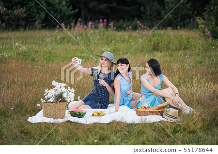 Three young women, in blue dresses and hats sit on a plaid and take pictures on a smartphone 55178644