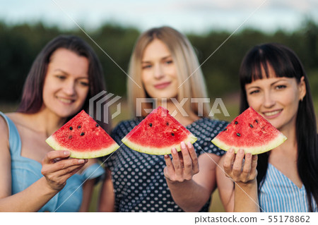 Three young attractive women friends in blue dresses at sunset are eating watermelon and smiling. 55178862