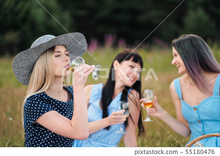 Three young women, blond, brunette and with dyed hair in blue dresses, and hats, sit on plaid and 55180476