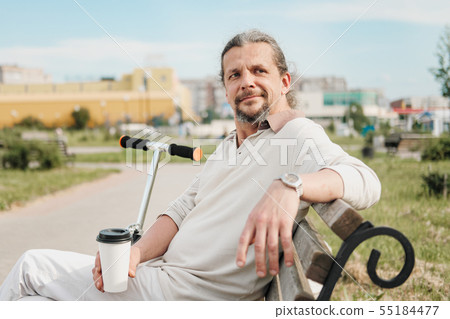 An attractive elderly 50-year-old man with long hair in the tail sits in a public garden on a bench 55184477