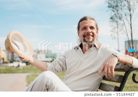 An attractive elderly 50-year-old man with long hair in the tail sits in a public garden on a bench 55184487