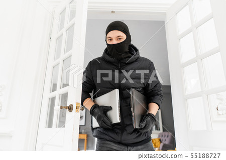 Thief looking at the camera with black balaclava stealing two modern expensive laptops. The burglar