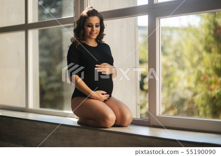 Pregnant moman sits in front of big window. Model dressed in black bodysuit. She put her hand on the Pregnant moman sits in front of big window. Model dressed in black bodysuit. She put her hand on the 55190090