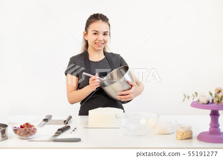 Attractive brunette girl pastry chef in apron whips cream cake in a metal bowl. Attractive brunette girl pastry chef in apron whips cream cake in a metal bowl. 55191277