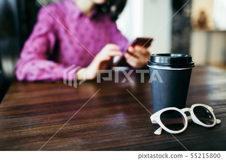 Young woman in the background scrolls her smartphone, cup of coffee at front. Young woman in the background scrolls her smartphone, cup of coffee at front. 55215800