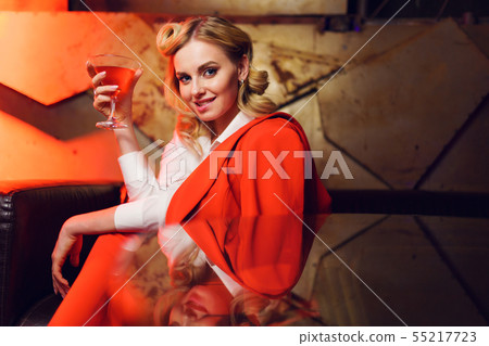 Image of young blonde in red jacket and trousers with cocktail in her hand in nightclub Image of young blonde in red jacket and trousers with cocktail in her hand in nightclub 55217723
