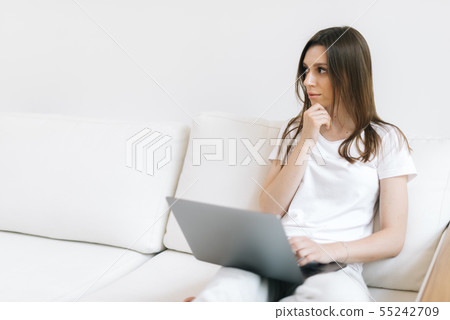 Close-up portrait of wondering girl looking away sitting at couch with laptop 55242709