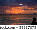 A tourist takes a picture of a sunset scene with red clouds and waves on the horizon on his mobile phone at a vacation spot. 55287875