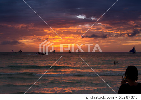 A tourist takes a picture of a sunset scene with red clouds and waves on the horizon on his mobile phone at a vacation spot. A tourist takes a picture of a sunset scene with red clouds and waves on the horizon on his mobile phone at a vacation spot. 55287875