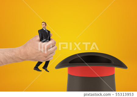 Man's hand holding tiny businessman and putting him in black top-hat on amber background with some 55313953