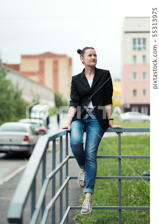 Young attractive woman in black jacket and blue jeans posing outdoor against background of building. 55313975