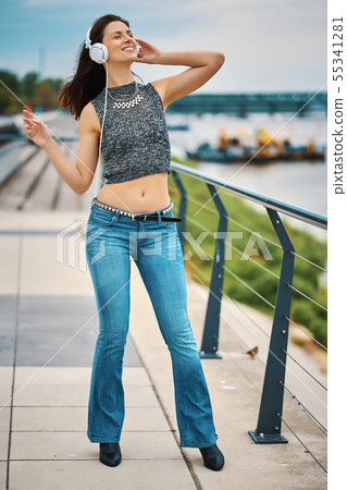 Beautiful woman with white headphones listens music while walking by the river in the city Beautiful woman with white headphones listens music while walking by the river in the city 55341281