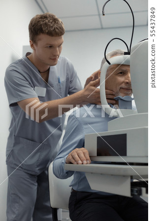 Dark-haired ophthalmologist assistant standing near patient Dark-haired ophthalmologist assistant standing near patient 55437849