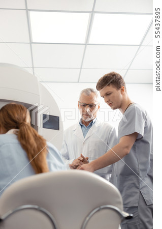 Dark-haired assistant of eye doctor helping him during examination Dark-haired assistant of eye doctor helping him during examination 55437895