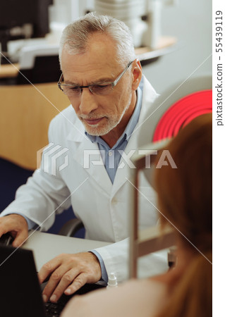 Grey-haired eye specialist looking at laptop while monitoring eye sight Grey-haired eye specialist looking at laptop while monitoring eye sight 55439119
