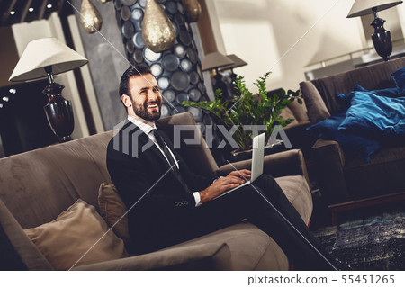Bearded young man smiling while sitting on the sofa with his modern device Bearded young man smiling while sitting on the sofa with his modern device 55451265