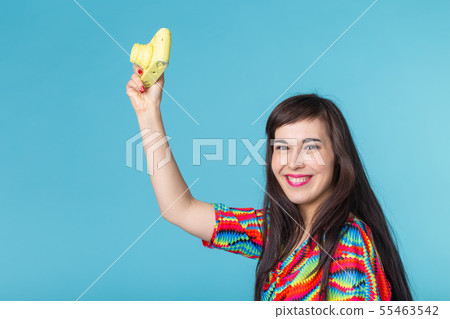 Smiling young woman is holding in her hands a yellow vintage camera model posing on a blue 55463542