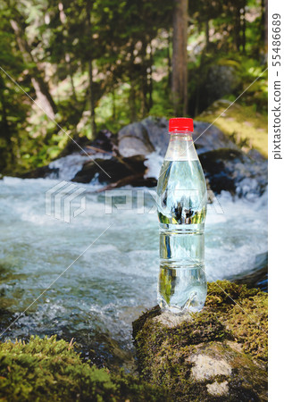 A plastic bottle with a red cap with fresh drinking water against a background of green forest and a 55486689