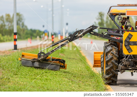 Tractor with a mechanical mower mowing grass on the side of the asphalt road. 55505347